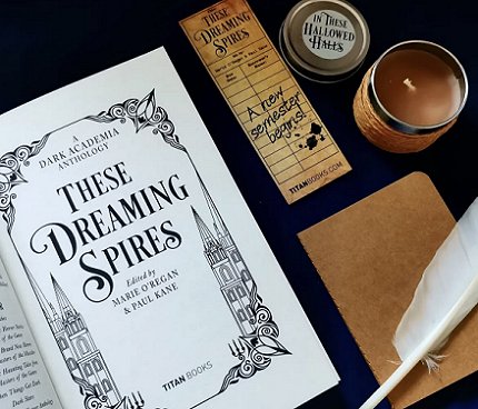 photograph of a copy of These Dreaming Spires, edited by Marie O'Regan and Paul Kane, open to show the title page, alongside a beige notebook and white feather quill pen, an In These Hallowed Halls jar candle and a Dreaming Spires bookmark