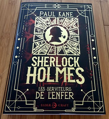 photograph showing a copy of the French Edition of Sherlock Holmes and the Servants of Hell (Sherlock Holmes, les serviteurs de l'enfer) by Paul Kane lying on a wooden surface