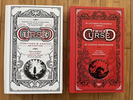 photograph showing the French editions of Cursed and Twice Cursed, edited by Marie O'Regan and Paul Kane, side by side against a wooden background