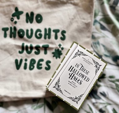 Image showing an open copy of In These Hallowed Halls, edited by Marie O'Regan and Paul Kane. Books is open to the title page, and is lying on a white cloth decorated with green leaves, and a beige bag with the words 'No Thoughts, Just Vibes'