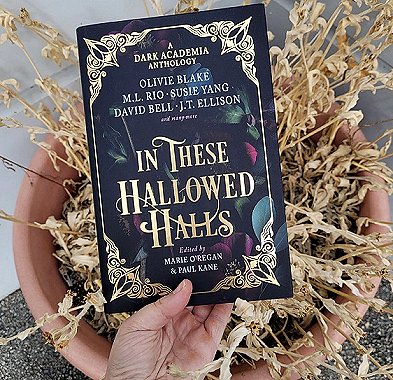 photograph of a hand holding a copy of In These Hallowed Halls, edited by Marie O'Regan and Paul Kane, over a terracotta planter containing a dead plant