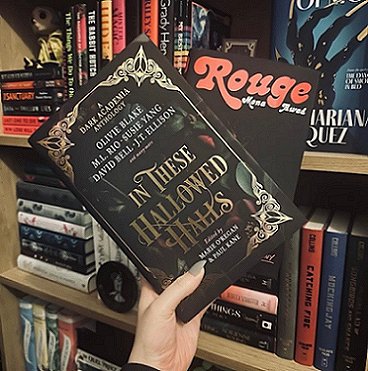 image of a woman's hand holding a copy of In These Hallowed Halls, edited by Marie O'Regan and Paul Kane, up against filled bookshelves