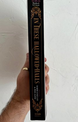 A man's hand holding a copy of In These Hallowed Halls, edited by Marie O'Regan and Paul Kane, so that the spine of the book faces out