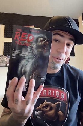 photograph of a man holding up a copy of The RED Trilogy by Paul Kane. Man is wearing a black baseball cap and a black t-shirt showing a wildcat and the word FLORIDA
