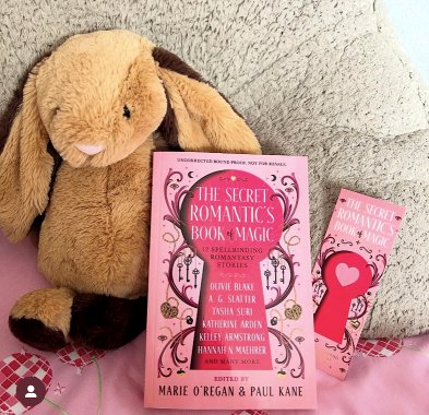 photograph of a copy of The Secret Romantic's Book of Magic, edited by Marie O'Regan and Paul Kane, lying beside a bookmark showing the book's cover. The book and bookmark are lying on a fleece cloth and pink bedspread alongside a brown toy rabbit