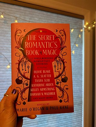 photograph of a woman's hand holding a copy of The Secret Romantic's Book of Magic, edited by Marie O'Regan and Paul Kane, against a window covered with a white blind decorated with lit fairy lights