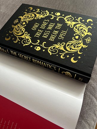 photograph showing the board cover inside the dustjacket of The Secret Romantic's Book of Magic, edited by Marie O'Regan and Paul Kane. The cover is black, decorated with gold embossed text reading ONLY TRUE LOVE'S KILL WILL BREAK THE SPELL..., surrounded by gold leaf decorations echoing the book cover