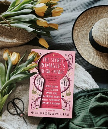 photograph of a copy of The Secret Romantic's Book of Magic, edited by Marie O'Regan and Paul Kane, on a cream fleece cloth, with a dark green scarf in the lower right corner, a straw hat with a black ribbon band and edging in the top right corner, and yellow tulips decorating the lefthand side