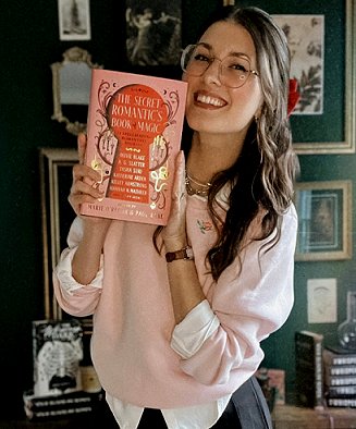 photograph of a smiling Kelly Andrew holding a copy of The Secret Romantic's Book of Magic, edited by Marie O'Regan and Paul Kane. Kelly is standing n front of a dark green 'gallery' wal, decorated with framed photographs and prints, with various books standing on a desk