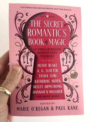 photograph of a man's hand holding a copy of the Secret Romantic's Book of Magic, edited by Marie O'Regan and Paul Kane, up against a white surface