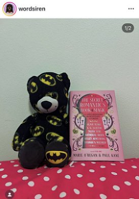 screenshot from @wordsiren: photograph of a copy of The Secret Romantic's Book of Magic, edited by Marie O'Regan and Paul Kane, standing on a pink cloth with white polka dots, beside a black teddy decorated with yellow and black Batman symbols