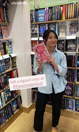 photograph of Olivie Blake standing in front of bookshelves, holding a copy of The Secret Romantic's Guide to Magic, edited by Marie O'Regan and Paul Kane. Text reads: left a signed copy at @bnsantamonica!