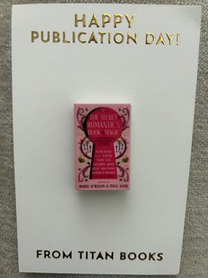 photograph showing a pin of the cover of The Secret Romantic's Book of Magic, edited by Marie O'Regan and Paul Kane, on a white card saying Happy Publication Day from Titan Books