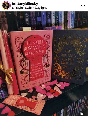 screenshot from @brittanykilinksy. Photograph of a copy of The Secret Romantic's Book of Magic, edited by Marie O'Regan and Paul Kane, standing alongside a copy without its dustjacket, to show the text: only true love's kiss will break the spell, standing in front of bookshelves. Alongside the books are two pink candles tied with gold ribbon, and pink heartshaped confetti is scattered in front of the books