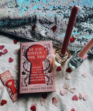 photograph of a copy of The Secret Romantic's Book of Magic, edited by Marie O'Regan and Paul Kane, lying on a white cloth covered with pink heartshaped confetti, alongside a Ssecret Romantic's bookmark and two pink candles in square glass candlesticks. At the top of the picture is a grey and blue cloth