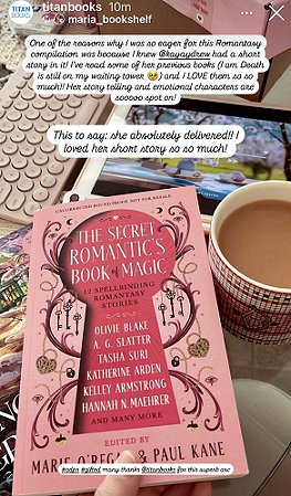 photograph of a copy of The Secret Romantic's Book of Magic, edited by Marie O'Regan and Paul Kane, being held above a computer mouse and a hardback book, with a cup of tea beside it. In the background is a computer keyboard, a tablet and at the top of the picture is a pink box