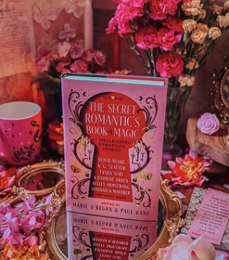 photograph showing a copy of The Secret Romantic's Book of Magic, edited by Marie O'Regan and Paul Kane, standing on a round gilt-edged mirror, surrounded by pink flowers, a Secret Romantic's bookmark, and a pink mug with black snakes on it