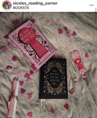 photograph of a copy of The Secret Romantic's Book of Magic, edited by Marie O'Regan - separated out so you see the pink dustjacket alongside the black bookcover, showing the text - only true love's kiss will break the spell... Alongside these are a Secret Romantic's bookmark and two candles, one pink, one white, tied with gold ribbon, and pink heartshaped confetti