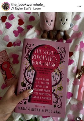 screenshot from @the_bookwormhole_. Photograph of a woman's hand holding a copy of The Secret Romantic's Book of Magic, edited by Marie O'Regan and Paul Kane, over a white cloth decorated with pink heartshaped confetti, alongside a Secret Romantic's bookmark on the left, a pink and a white candle tied with gold ribbon on the right, and pink and white smiling felt toys above